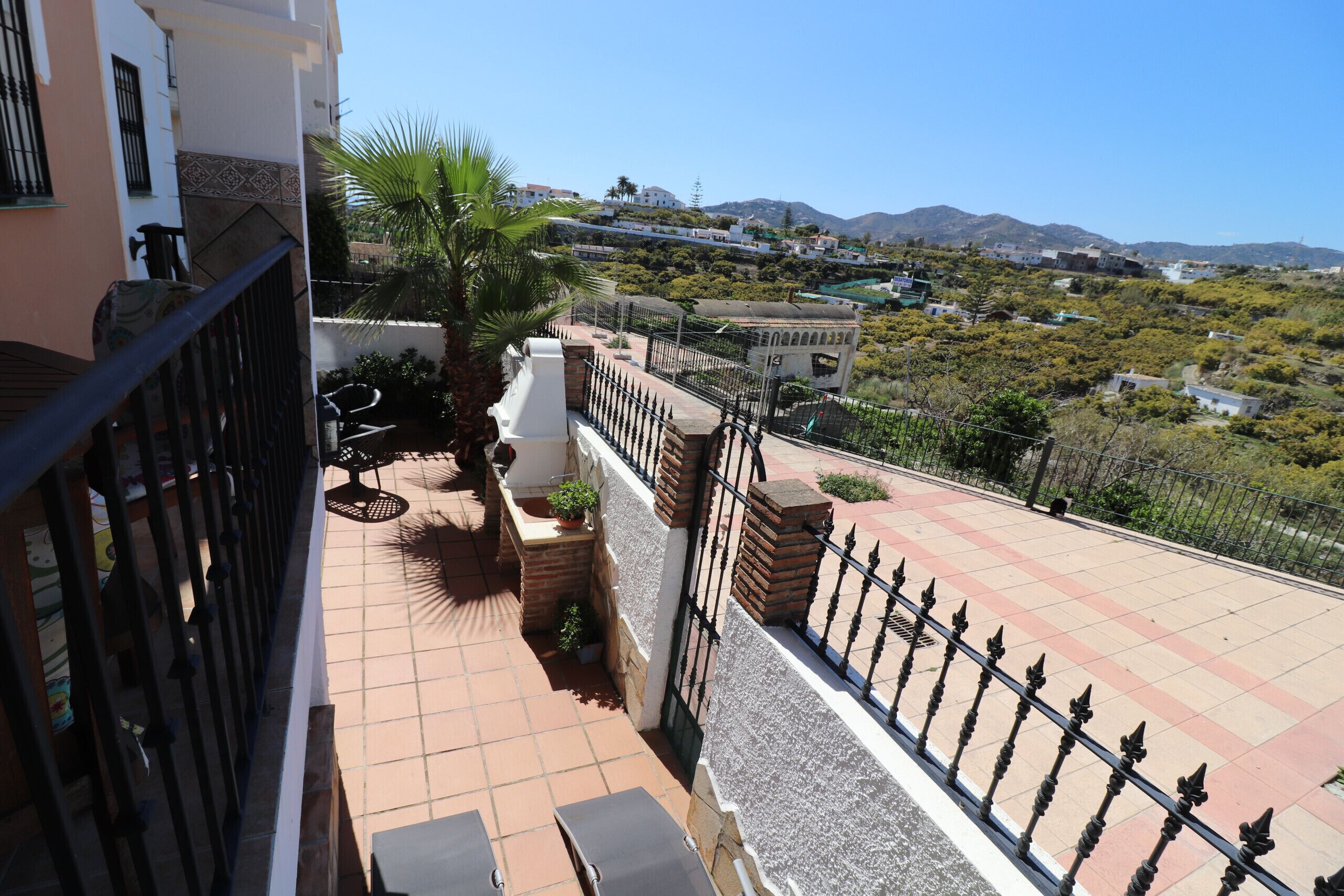 A terrace with a tiled floor, a black metal railing, and a view of a hillside with buildings and greenery.