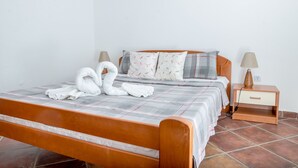A neatly made bed with a checkered bedspread, two pillows with a leaf pattern, and a pair of white towels arranged in a swan shape. There is a wooden nightstand with a lamp and a drawer.