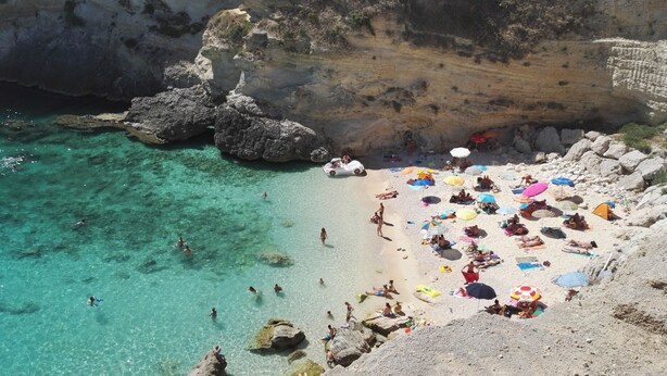 Una playa con agua turquesa cristalina, formaciones rocosas y gente tomando el sol y nadando.