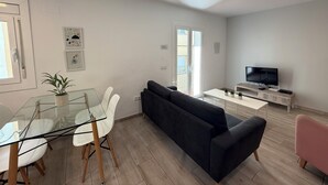 A modern living room with a glass dining table, white chairs, a black sofa, a TV on a wooden stand, and a sliding glass door leading to a balcony.