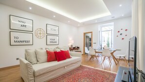 A modern living room with a sofa, a coffee table, and a dining area with chairs. There are framed motivational quotes on the wall and a large window providing natural light. - The Dreams House with Green Patio (Lisboa)