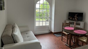 A room with a white sofa, a dining table with chairs, a television on a stand, and a large arched window.