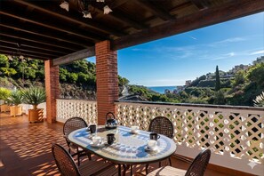 A covered outdoor seating area with a table set for two, surrounded by chairs, and overlooking a scenic hillside. - 1037 Villa Alabarce (Nerja)