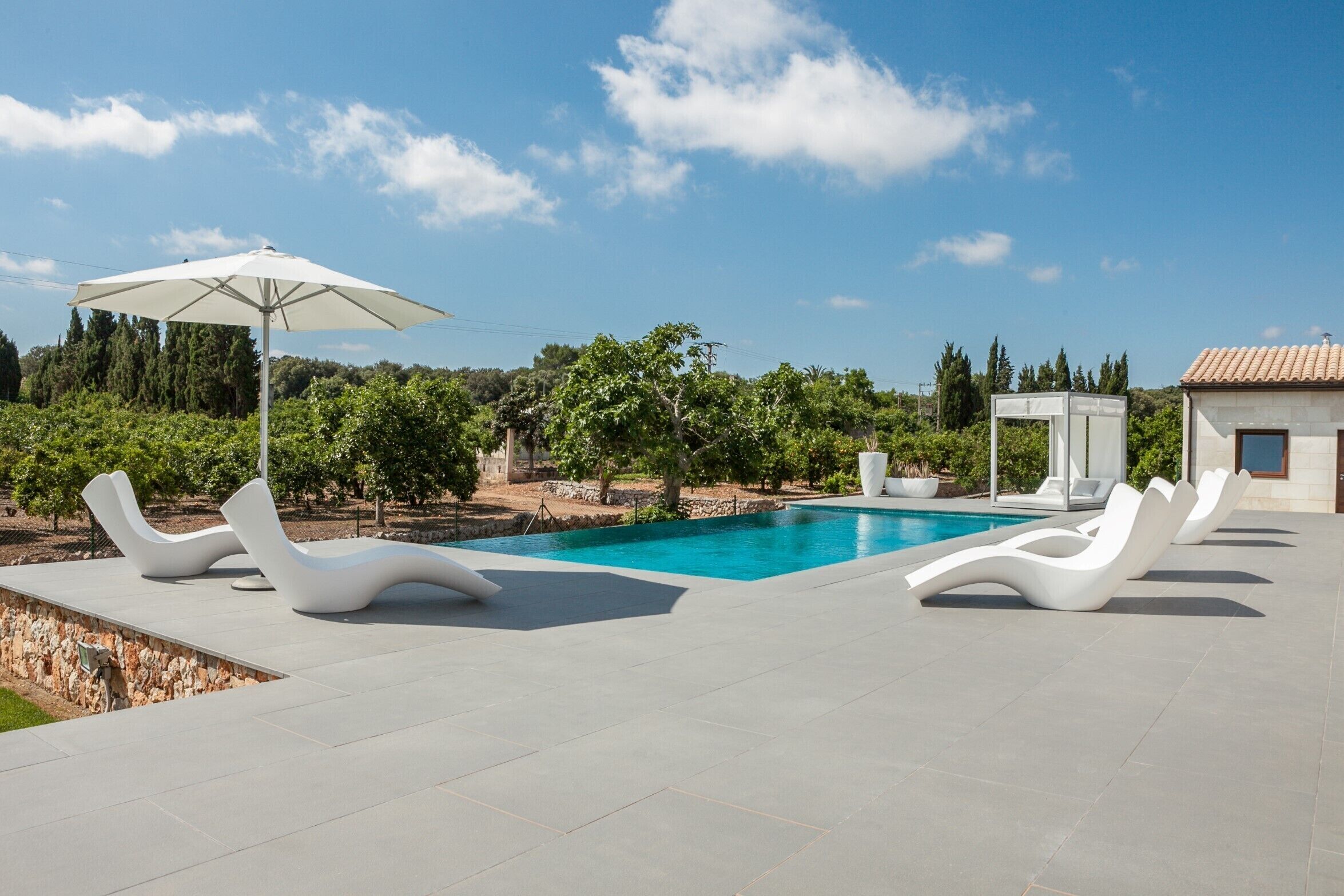 Un área de piscina moderna al aire libre con sillones blancos y una pérgola con sombrilla.