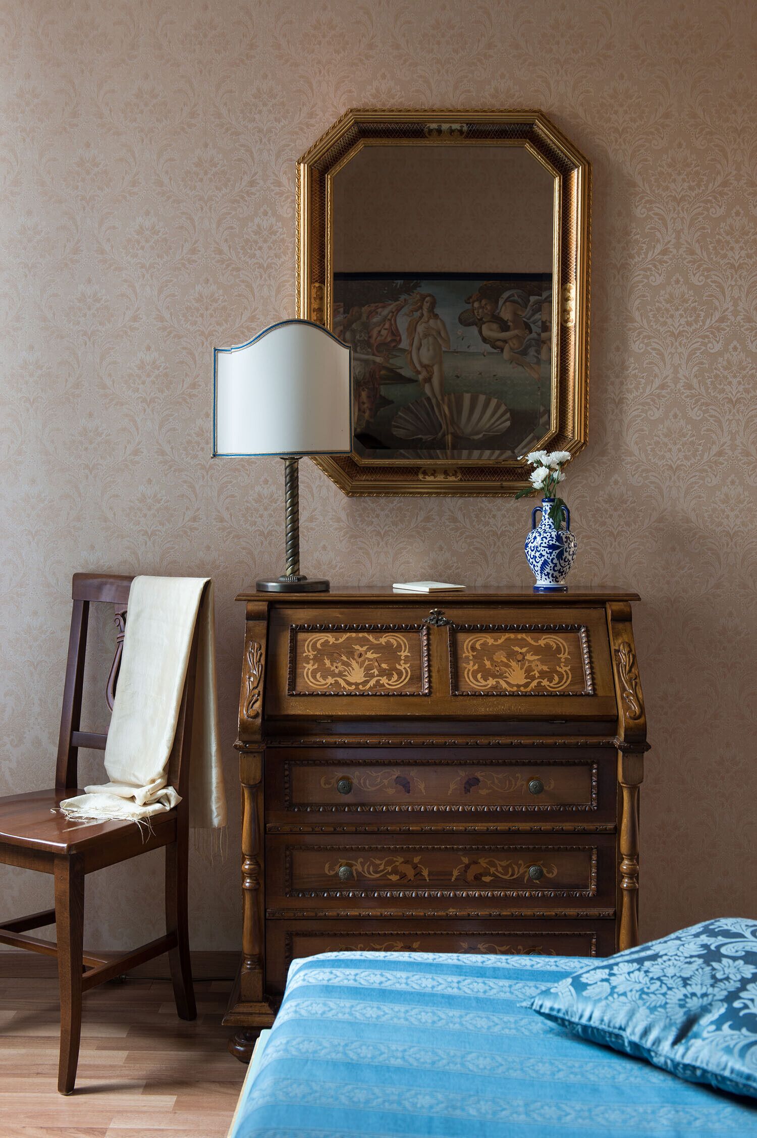 Une pièce comprenant une commode en bois, une chaise avec une serviette blanche, une couvre-lit bleue et un miroir orné d’un tableau.