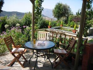 A patio with a round table and two wooden chairs, surrounded by potted plants and a view of hills. - Garden apartment in the heart of the countryside, close to local villages. (Pórtugos)