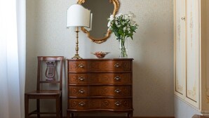 Ett rum med en byrå i trä, en spegel, en lampa, en vas med blommor, en stol, ett bord och en mönstrad matta.