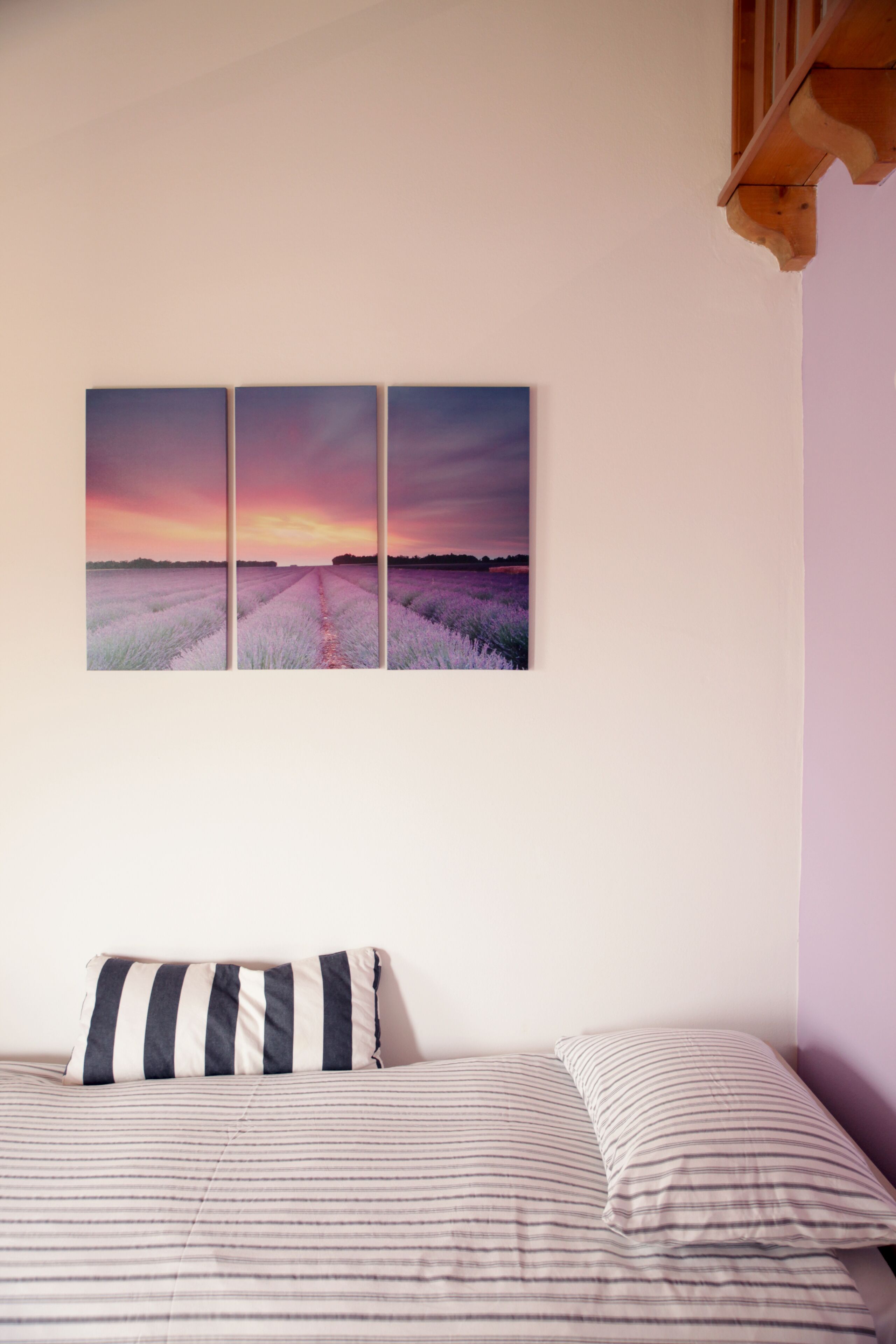 Ein Schlafzimmer mit einem Bett, einem gestreiften Kissen und einem Wandbild einer Lavendelfeld-Szene bei Sonnenuntergang.