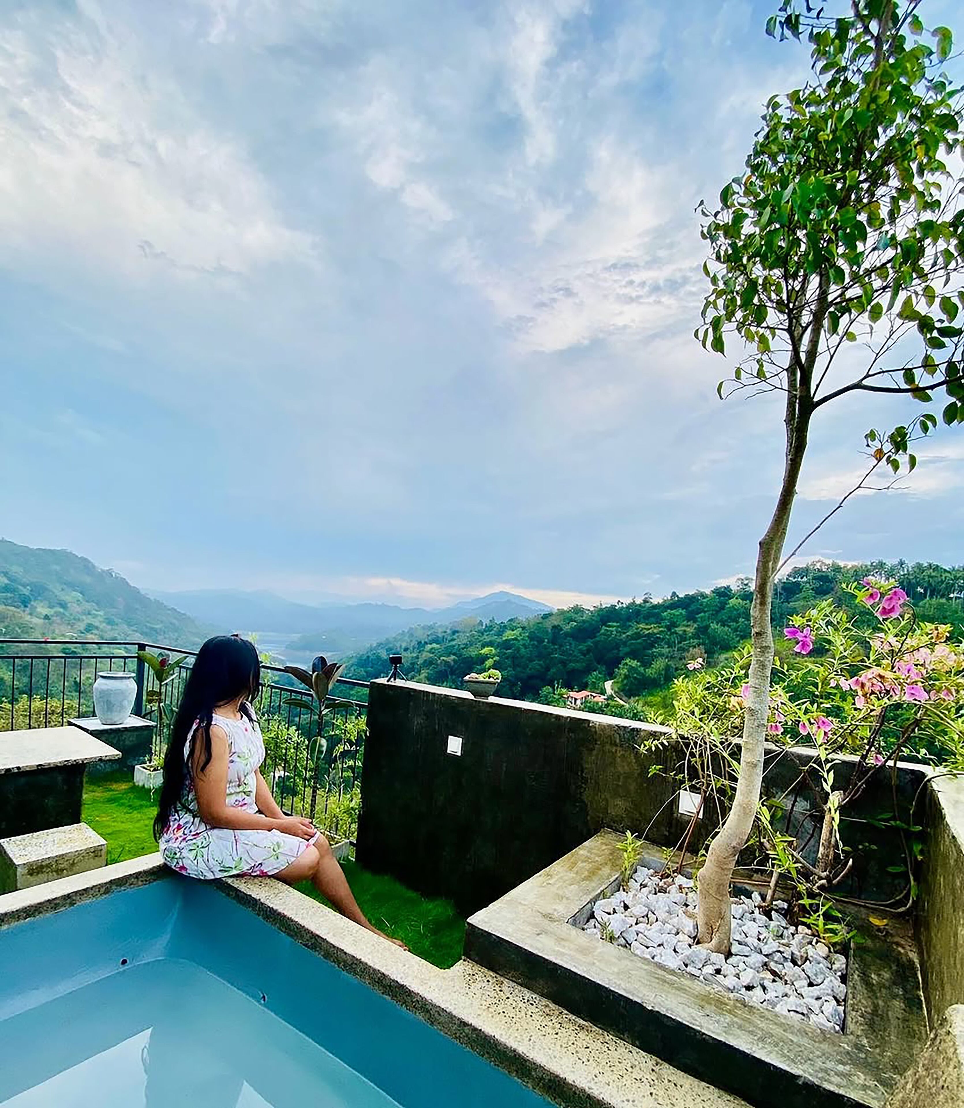 A woman sitting by a pool with a view of mountains and greenery.