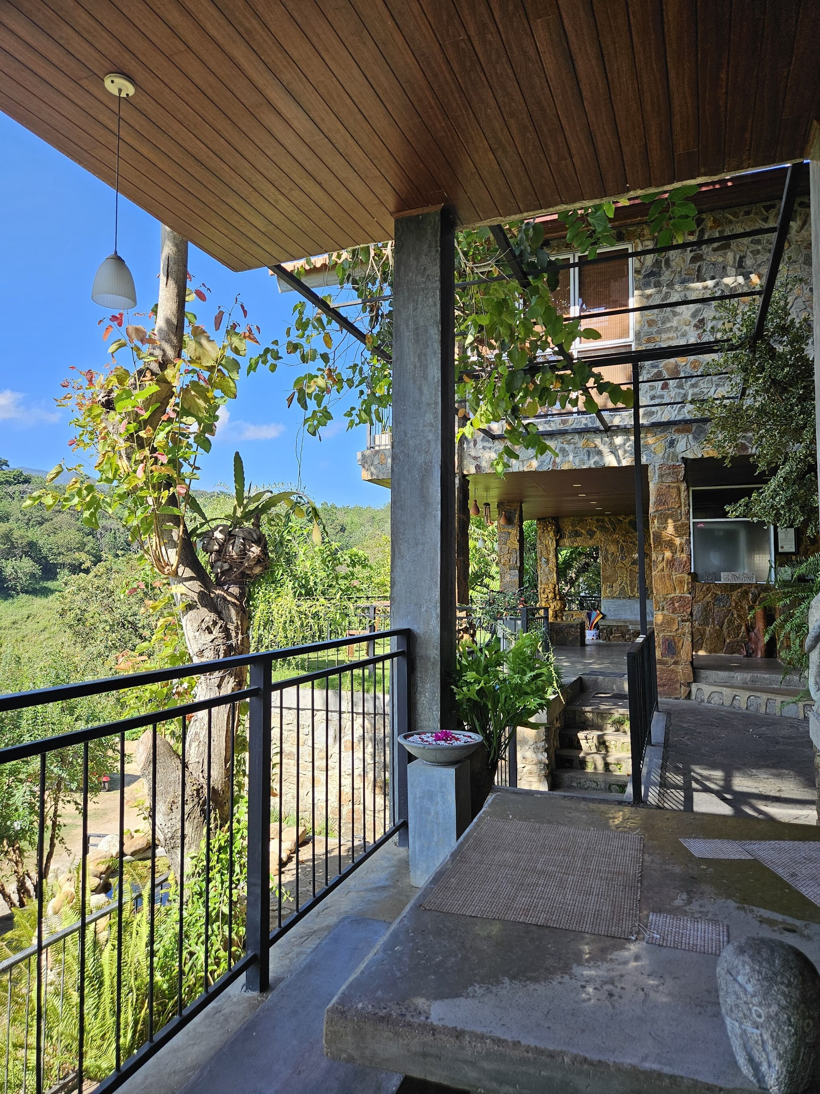 A covered outdoor area with a stone wall, a metal railing, and a view of a lush landscape.