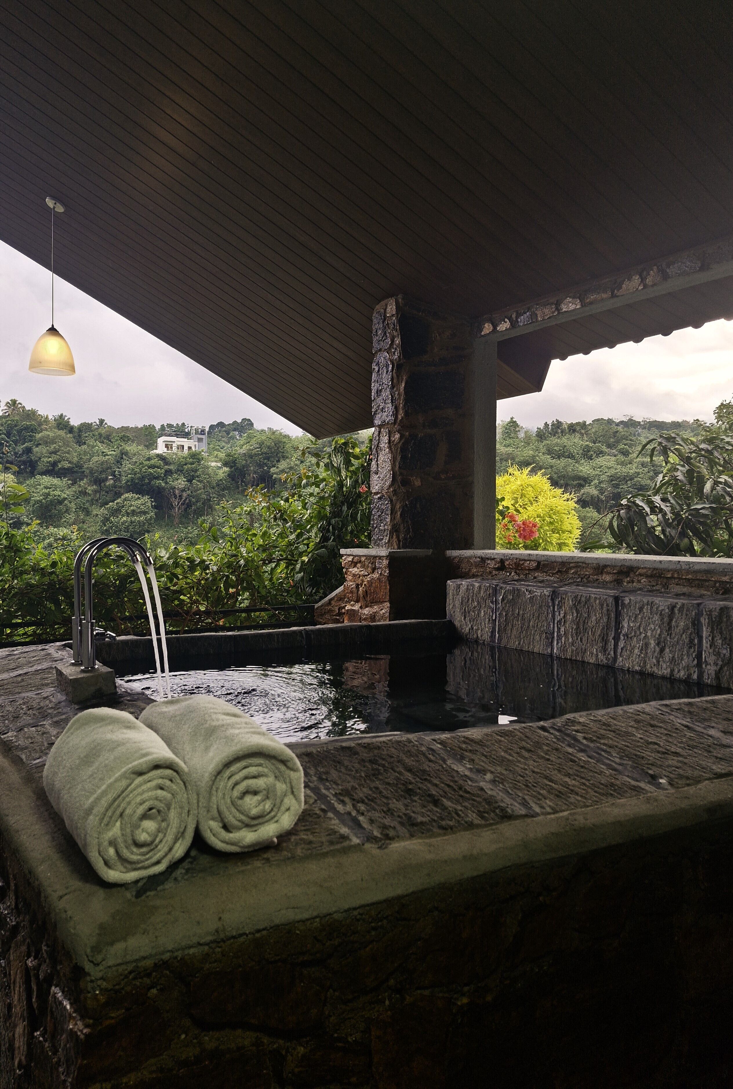 A stone hot tub with a waterfall feature, surrounded by lush greenery and a stone bench with towels.