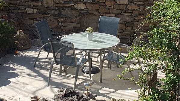 An outdoor patio with a round glass table surrounded by metal chairs, set against a stone wall.