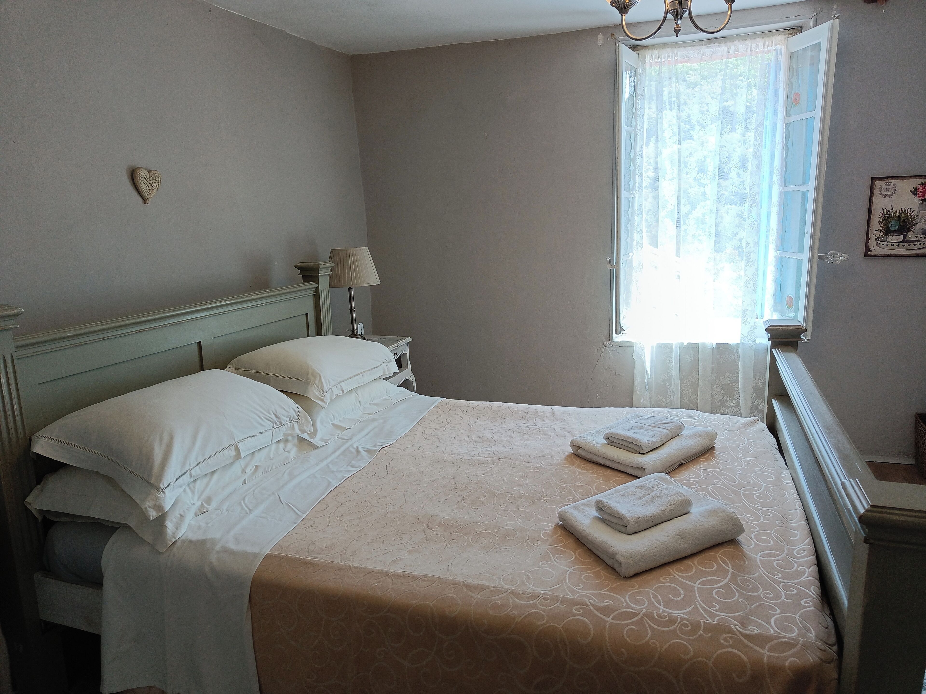 A neatly made bed with white linens and a beige patterned bedspread, a bedside lamp, and a window with sheer curtains.
