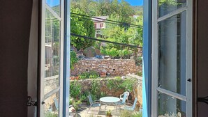 Vista per una finestra oberta d'un pati amb mur de pedra, mobles d'exterior i un paisatge de muntanya de fons.