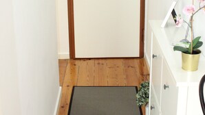 A hallway with a wooden floor, a mat, a white door with a green sign above it, a white dresser with a mirror, and a potted orchid.