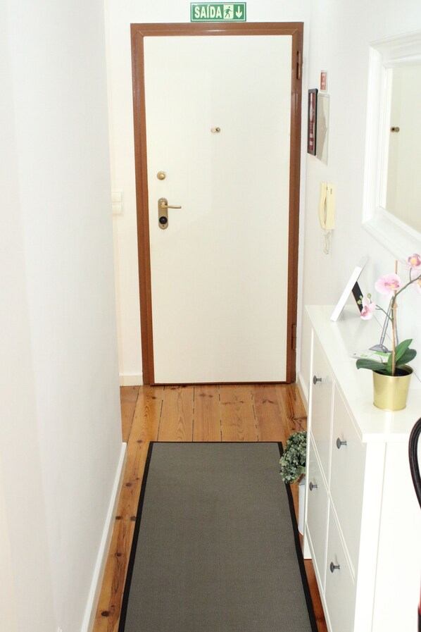A hallway with a wooden floor, a mat, a white door with a green sign above it, a white dresser with a mirror, and a potted orchid.