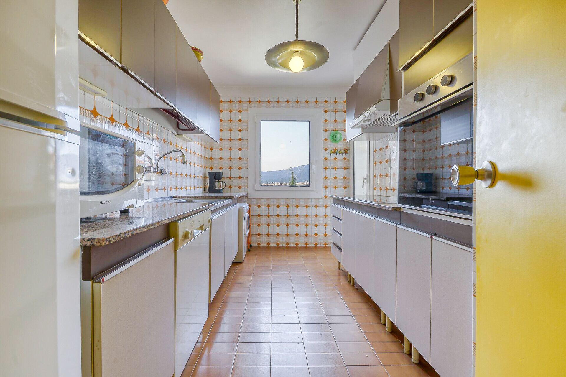 Een smalle keuken met witte kastjes, een raam en uitzicht op de omgeving.