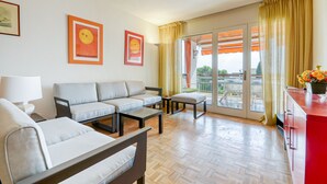 A modern living room with a sofa, armchair, and coffee table, a balcony with yellow curtains, and a view of the outdoors.