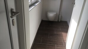 Un pasillo estrecho con suelo de madera oscura, una puerta blanca con cerradura, una ventana con persiana blanca y un inodoro blanco con palanca.