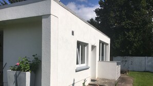 Un edificio moderno con fachada blanca, un pequeño jardín con una flor rosa y un sendero pavimentado que conduce a un banco de madera.