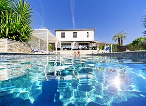 Ein Swimmingpool mit klarem, blauem Wasser, Liegestühlen und ein Gebäude mit einem Kachel­dach im Hintergrund.