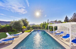 A swimming pool with lounge chairs, surrounded by greenery and a clear sky.