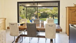 A dining area with a wooden table and four chairs, a view of a pool and greenery, and a kitchen with a refrigerator and wooden cabinets. - Villa with pool for discerning connoisseurs (Bagnols en foret)