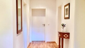 A hallway with wooden flooring, white walls, and a door at the end. There are two framed pictures on the walls and a small table with a vase on the right.