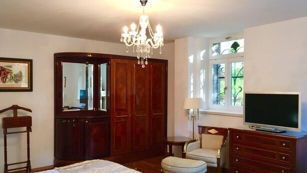 A bedroom with a bed, a dresser, a television, a chair, a mirror, and a chandelier.