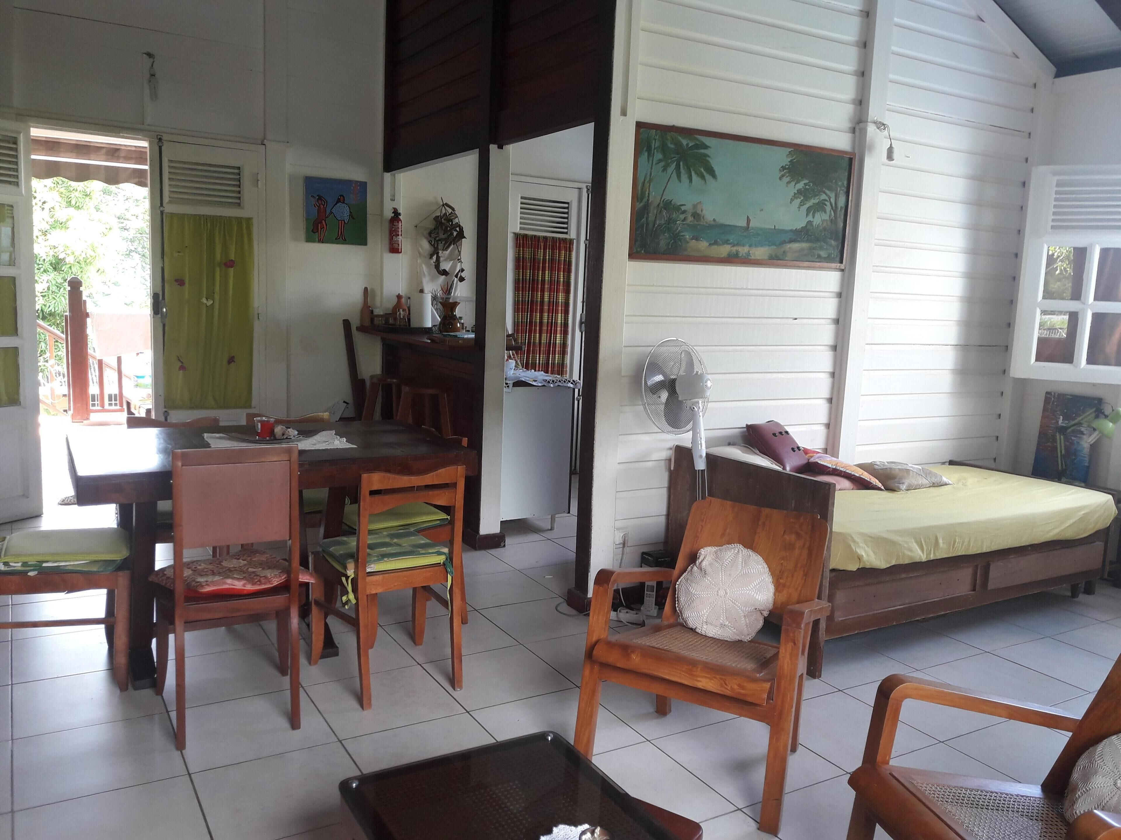 Una habitación con una mesa de comedor de madera, sillas, una cama con colchón amarillo, un ventilador y un cuadro de una escena de playa.