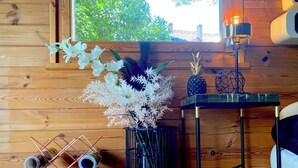A wooden interior with a wooden wall, a window with a view of greenery, a table with a pineapple, a lamp, and a vase with white flowers. - House with garden in the center of Berlin for up to 3 people (Berlin)