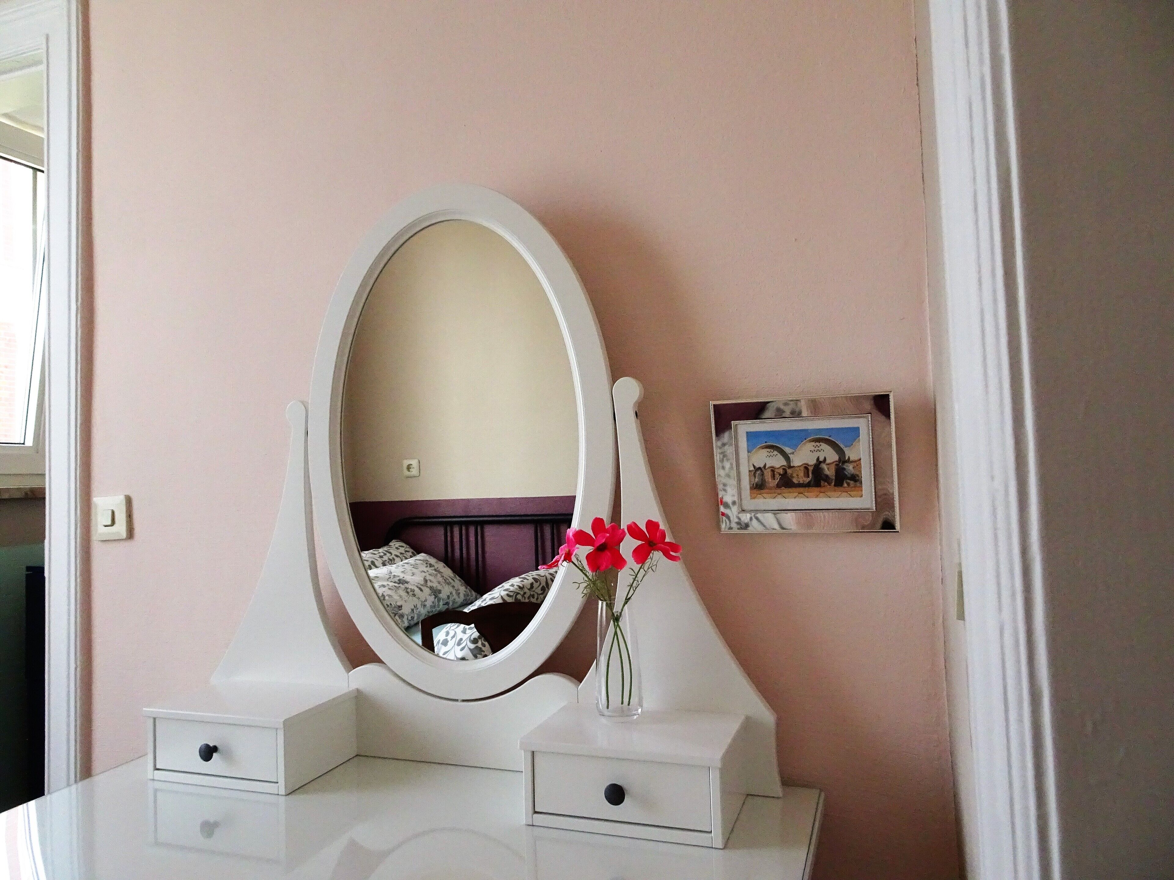 Een witte kaptafel met een ronde spiegel, een klein vaasje met bloemen en een fotolijst aan de muur.