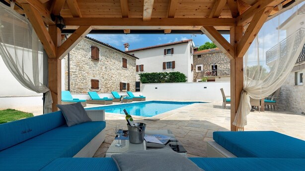 A covered outdoor seating area with blue sofas, a pool, and stone buildings in the background. - Villa Loki- your exclusive Holiday Home in Istria (Lašići, Vizinada)