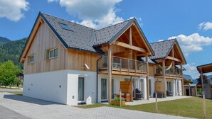 Una casa moderna de madera con balcón, un área de estar al aire libre techada y un camino de entrada pavimentado.