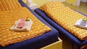 A hotel room with two single beds, each with a patterned bedspread and pillows. There is a bedside table with a lamp and a small box, possibly for toiletries. The room has a simple layout with a plain wall and a door leading to another room. - Small apartment central location Fulda Rhön Vogelsberg Hesse 2 pers. (Künzell)
