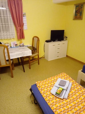 A small room with a bed, a desk, a chair, a television on a cabinet, and a window with curtains.