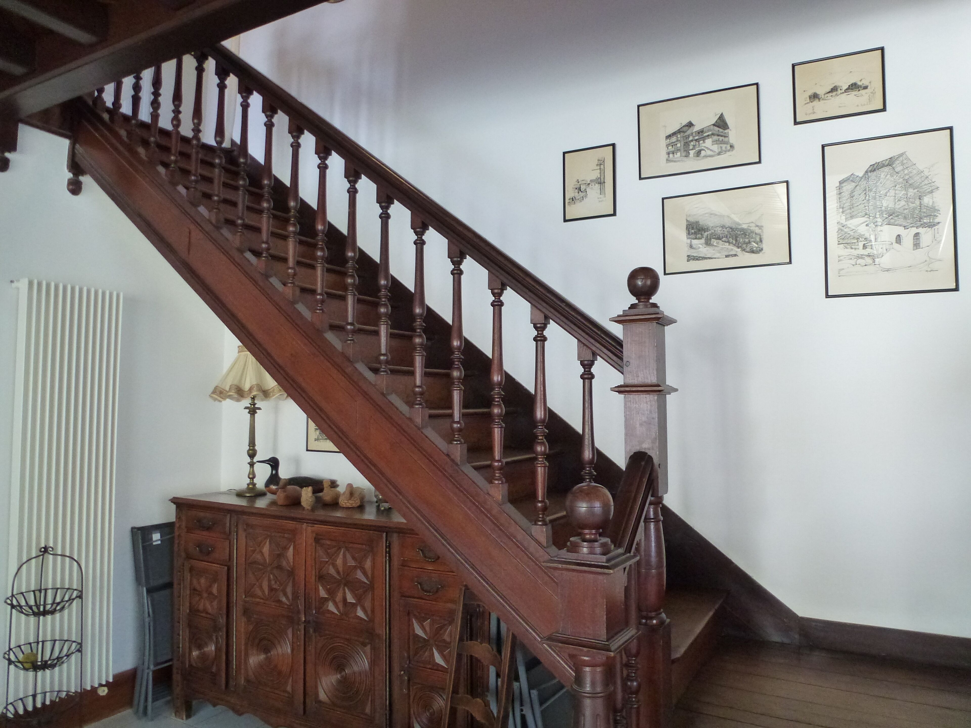 Una escalera de madera con barandilla, un radiador, un armario con adornos tallados y cuadros enmarcados en la pared.