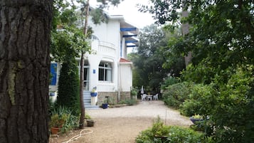Un edificio blanco con balcón azul, rodeado de árboles y un camino de grava.