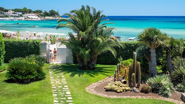 Un jardin en bord de mer, avec un sentier menant à un point de vue sur la plage, agrémenté de cactus et de palmiers.