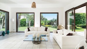 Un salon spacieux avec un canapé blanc, une table basse en verre et de grandes fenêtres donnant sur un jardin luxuriant.