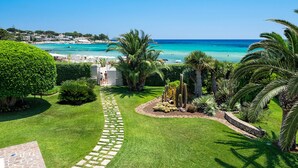 Un jardin bien entretenu, avec un chemin de pierre menant à un bâtiment blanc, entouré d’une végétation luxuriante et de palmiers, et offrant une vue sur une plage aux eaux turquoise et cristallines.