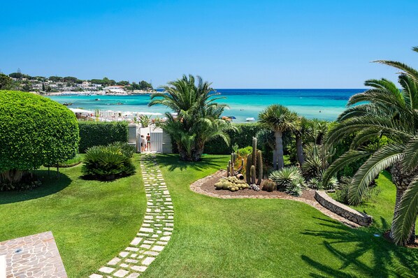 A well-maintained garden with a stone pathway leading to a white building, surrounded by lush greenery and palm trees, overlooking a beach with clear turquoise waters.