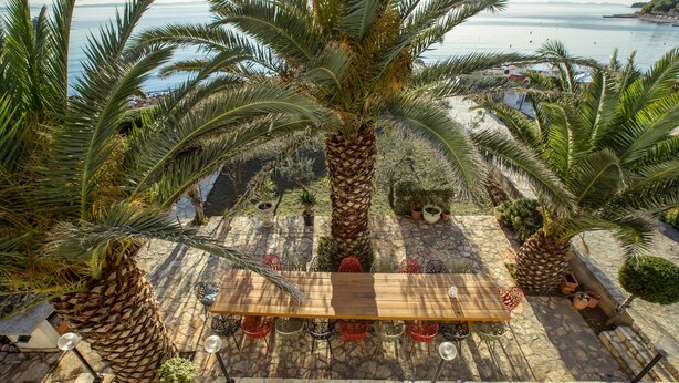 Een terras met een houten tafel en stoelen, omringd door palmbomen en plantenbakken, met uitzicht op een wateroppervlak.