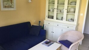 A living room with a dark blue sofa, a white table with magazines, a wicker chair, and a white cabinet with glass doors containing dishes and glasses. - House 2 bedrooms Pierre et Vacances Port Bourgenay Domaine du Moulin (Talmont -Saint-Hilaire)