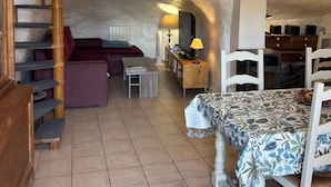 A rustic interior with a wooden staircase, a dining table with a floral tablecloth, a sofa, and a kitchen area with a clock. - House "Ciboulette (Mirabel-aux-Baronnies)
