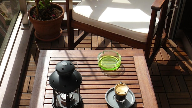 A wooden table with a coffee cup and a green ashtray, a wooden chair with a white cushion, and a potted plant on a balcony with a view of a building. - Beautiful studio Beautiful view (Strasbourg)