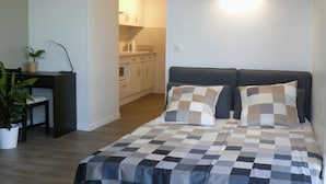 A bedroom with a checkered bedspread, two pillows, a desk with a chair, a potted plant, and a kitchenette in the background. - Beautiful studio Beautiful view (Strasbourg)