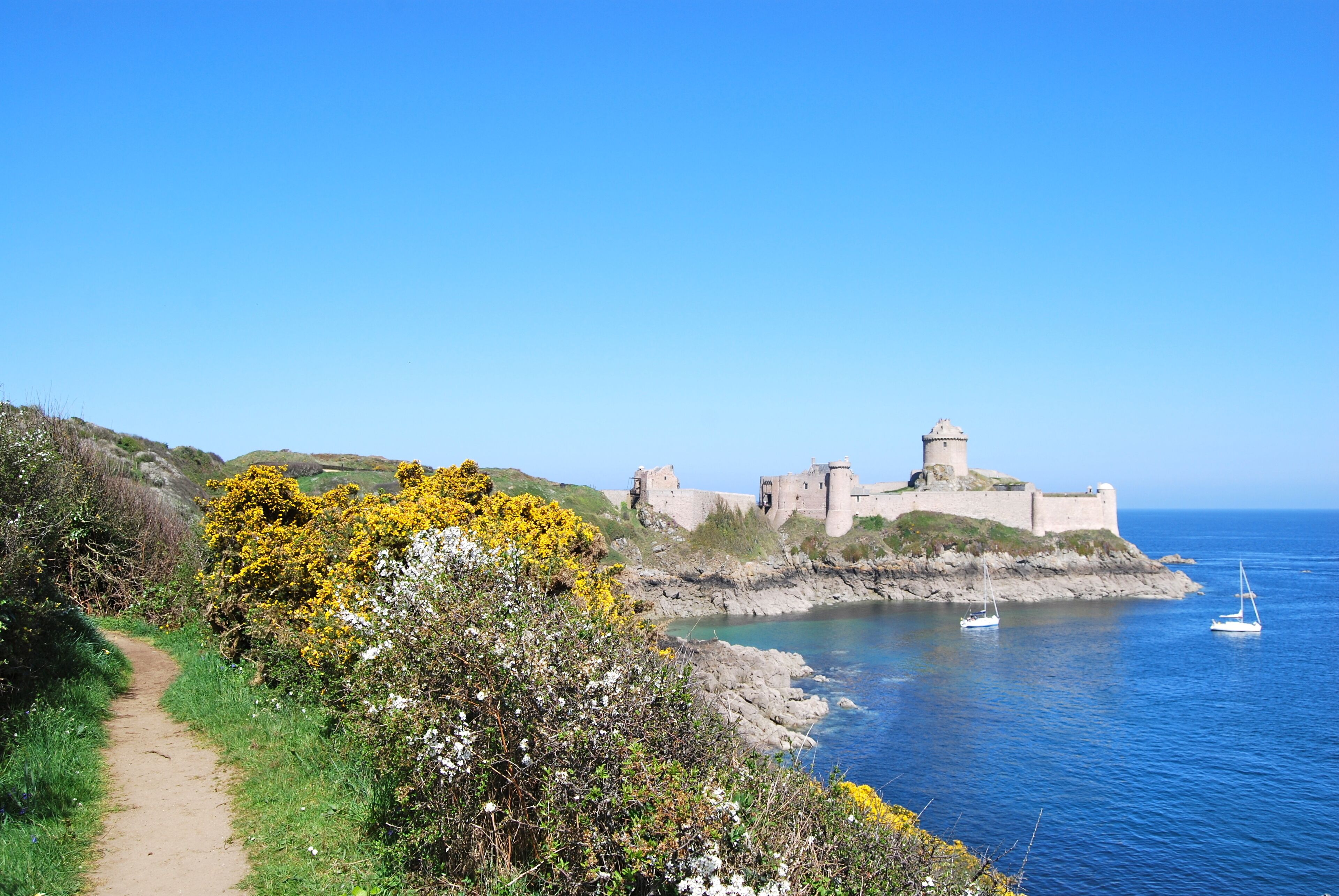 A coastal path leading to a historic fortification with a clear view of the sea and a sailboat.