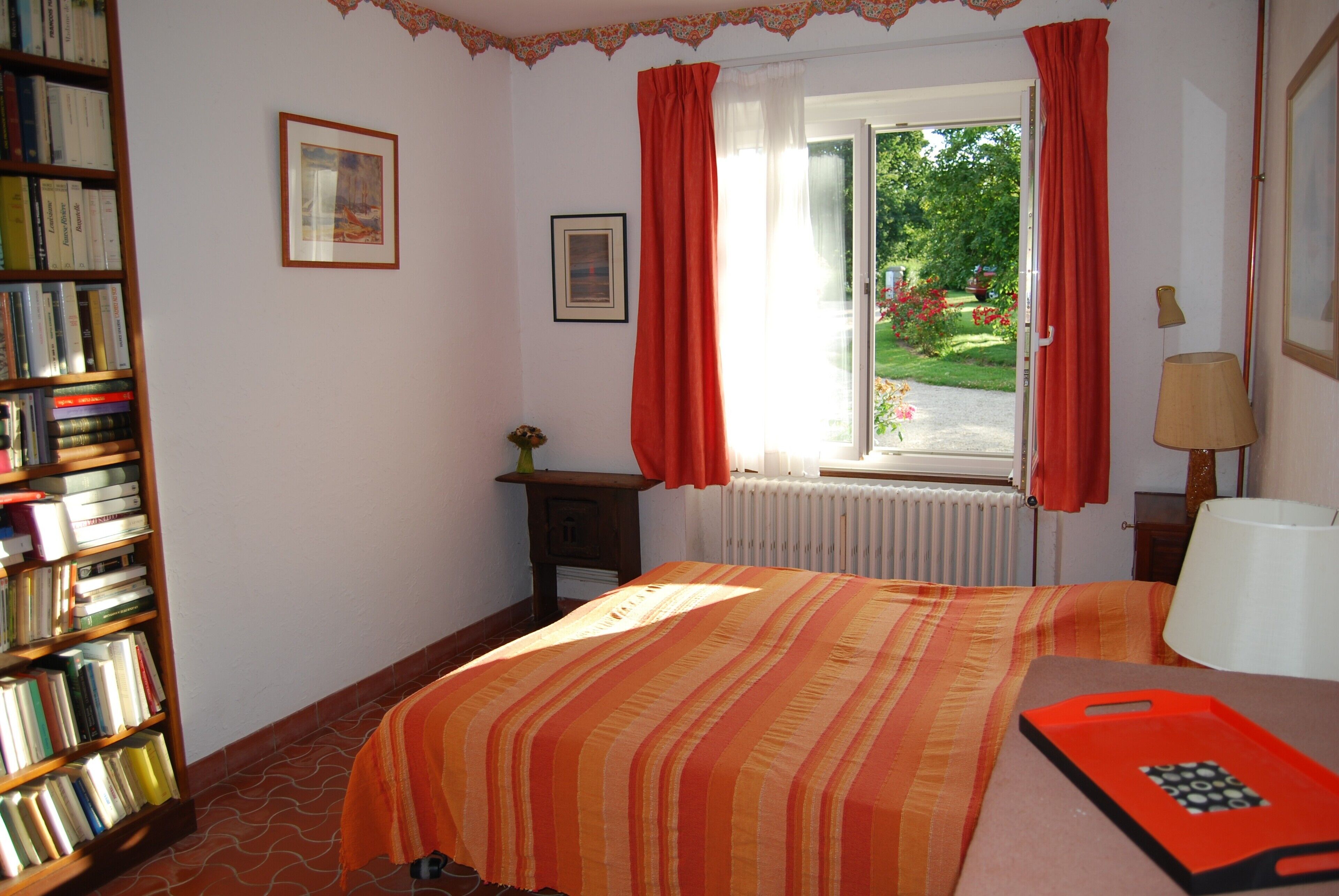A bedroom with a bed, a bookshelf, a window with curtains, a radiator, and a bedside table with a lamp.