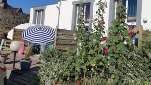 A white building with a red roof, a chimney, and a window with a red curtain. There is a blue and white striped umbrella and a potted plant with pink flowers.